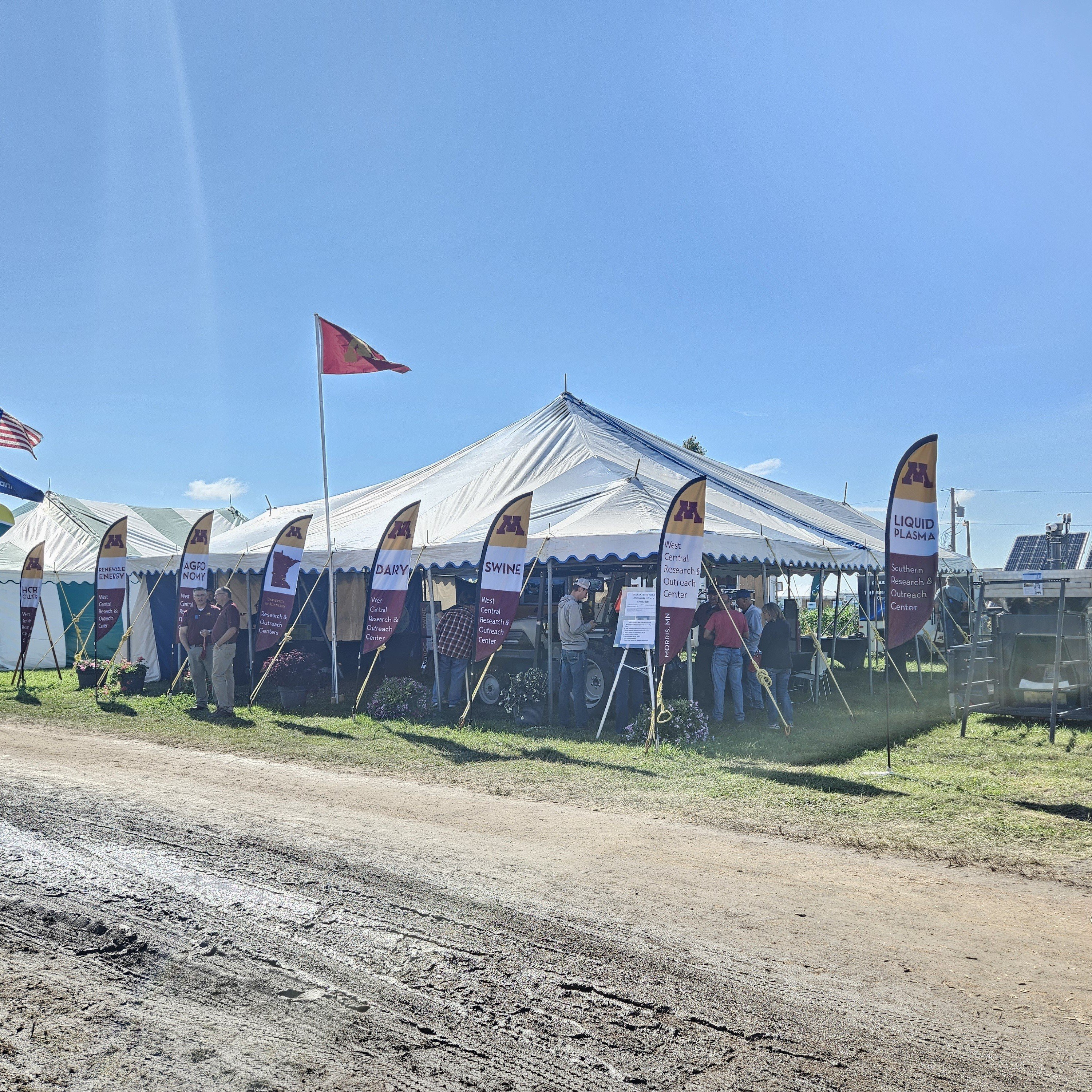 A festival tent with branded UMN feather flags waving out front. A muddy path fills the foreground.