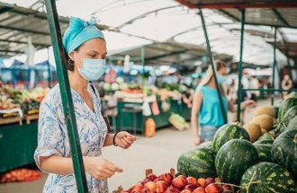 A woman with a head scarf and a blue face mask looks at watermelons and peaches at a farmers market.