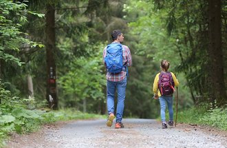 An adult and child walk together on a wooded trail.